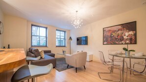 A modern living room with a wooden dining table, a grey sofa, a glass coffee table, and a large painting on the wall. - Hampden Apartments - The Catherine (Windsor)