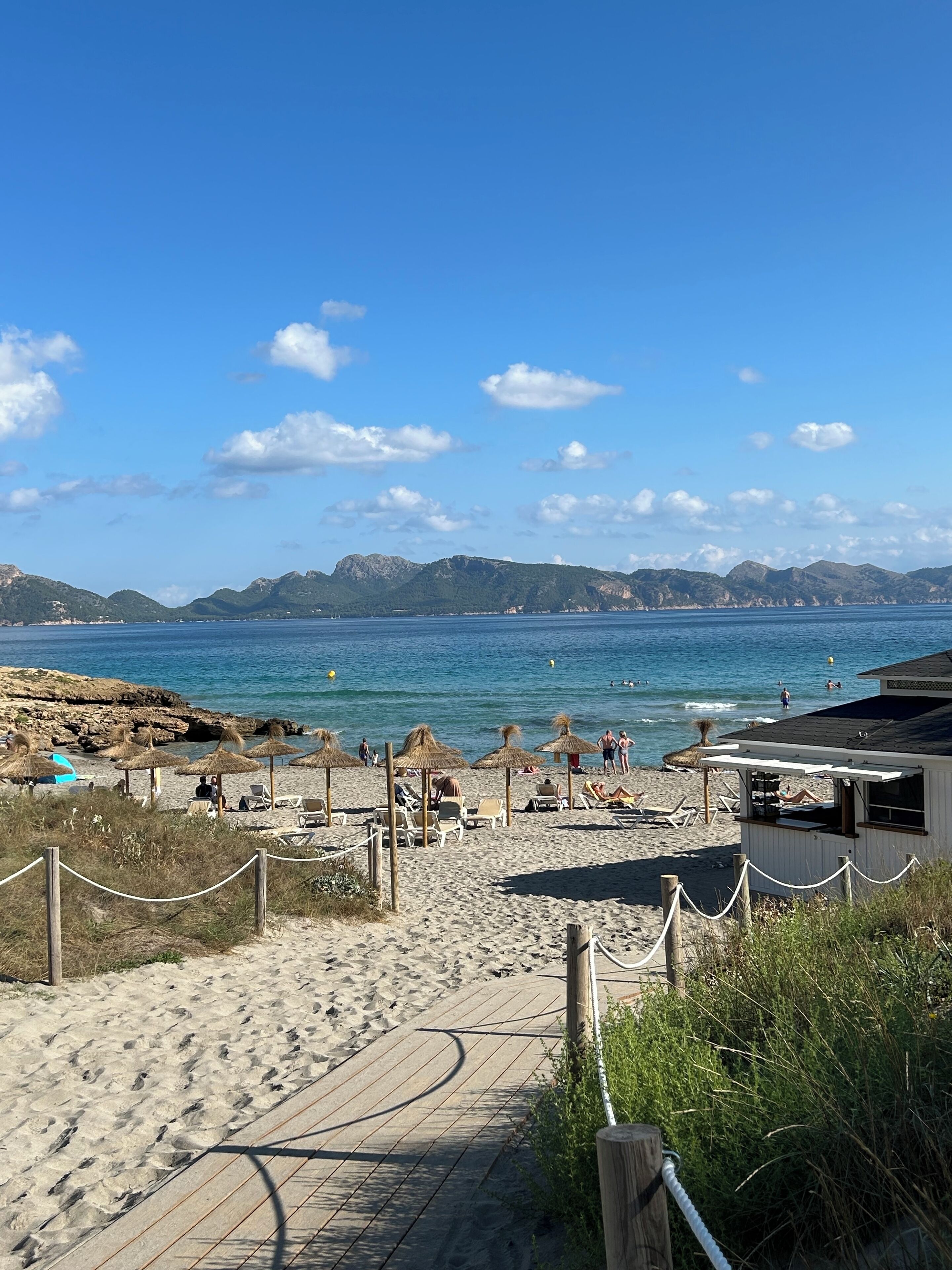 A beach with a wooden boardwalk leading to the shore, thatched umbrellas, and a small building with a dark roof.