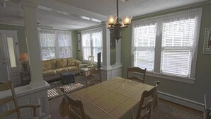 Uma sala de jantar com mesa, cadeiras e um candelabro. Há um sofá e uma janela com persianas ao fundo.