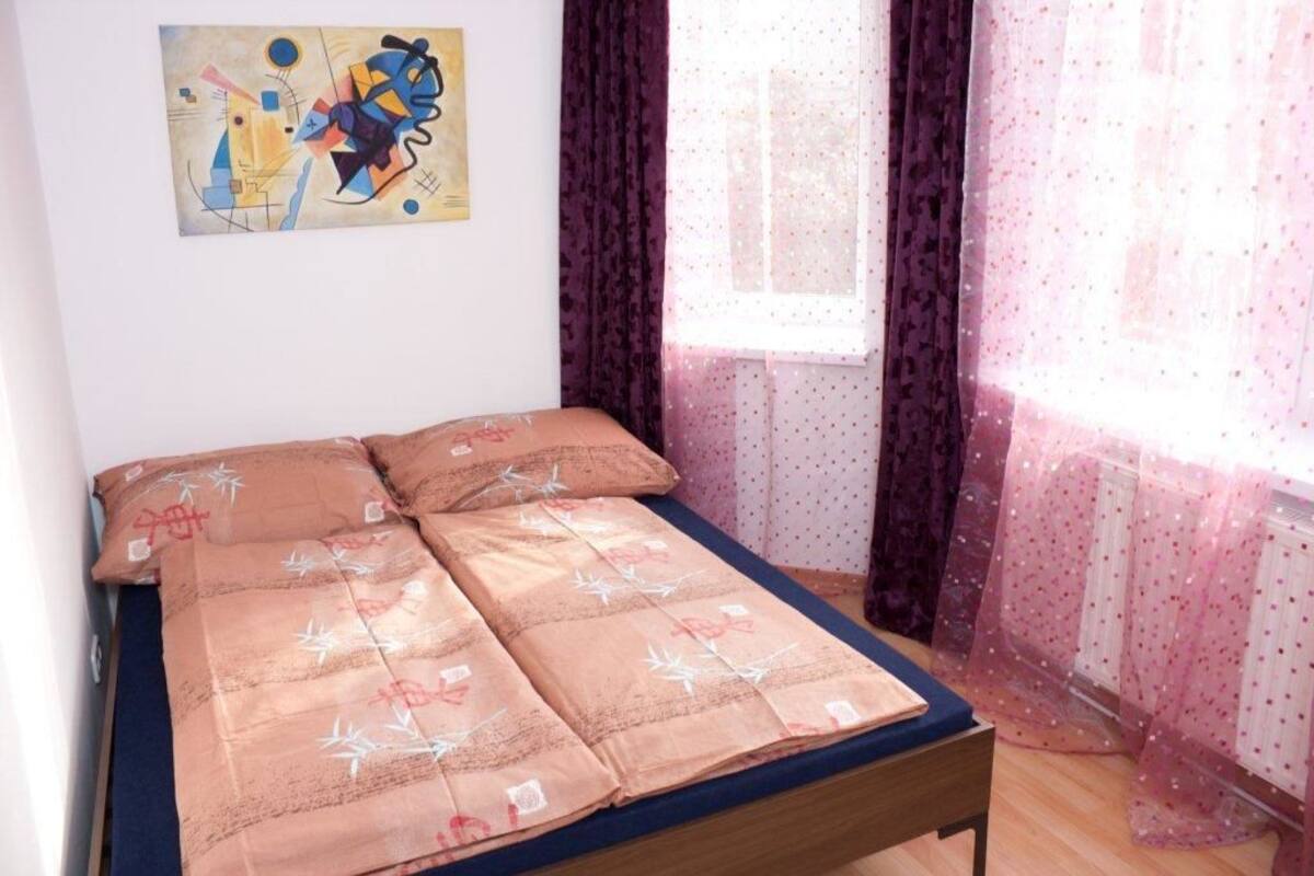 A bed with a floral bedspread, a window with sheer curtains, and a painting on the wall.