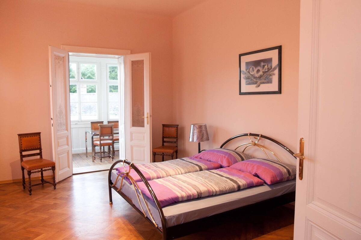 A bedroom with a bed, a chair, a desk, and a door leading to a balcony.