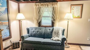 Un salón acogedor con un sofá azul, dos lámparas, una cita enmarcada en la pared y una ventana con cortinas traslúcidas.