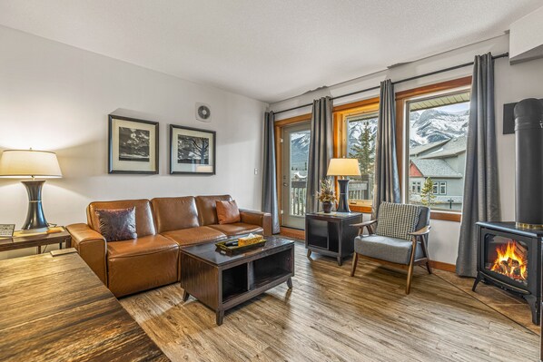 Ein gemütliches Wohnzimmer mit Ledersofa, Couchtisch, Kamin und Blick auf Berge durch große Fenster.