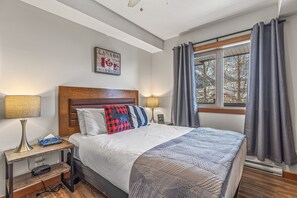 Ein ordentlich eingerichtetes Schlafzimmer mit einem großen Bett, einem Holz-Kopfteil, einem Nachttisch mit Lampe und einem Fenster mit Vorhängen.