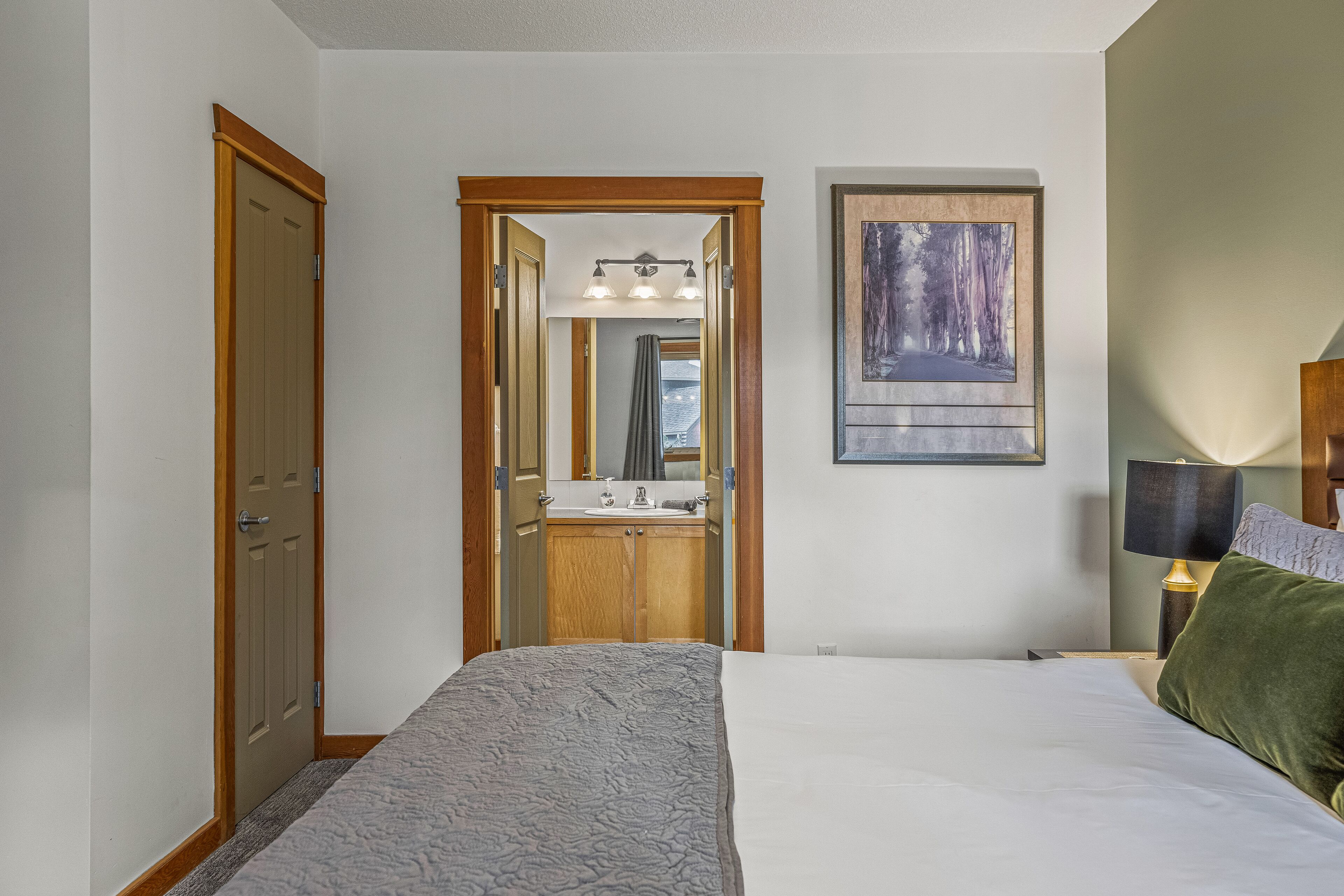 Un dormitorio con cama, mesita de noche, lámpara, un cuadro, baño con lavabo y espejo, y una puerta.
