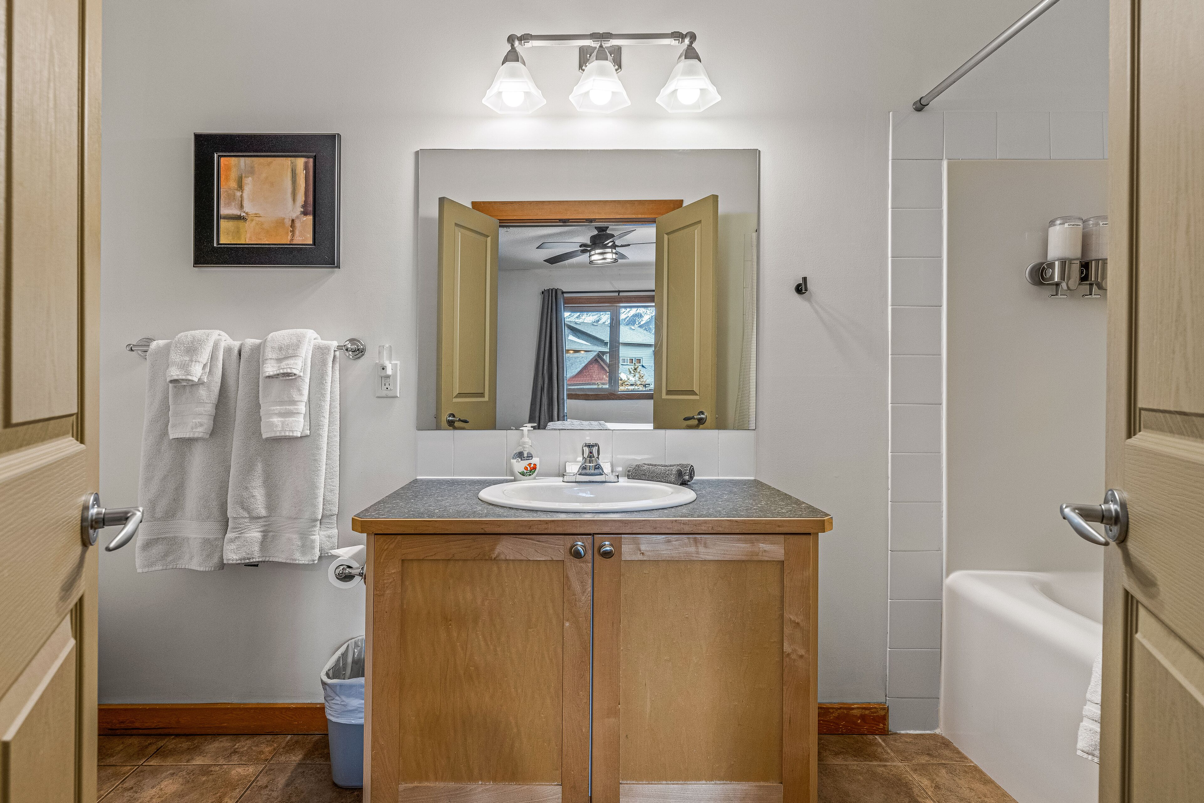 A bathroom with a wooden vanity, a white sink, a mirror with lights above, a towel rack, and a shower area.