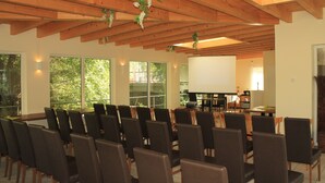 A conference room with rows of chairs, a projector screen, and large windows with a view of greenery. - Land Gut Hotel Weisser Schwan (Zossen)
