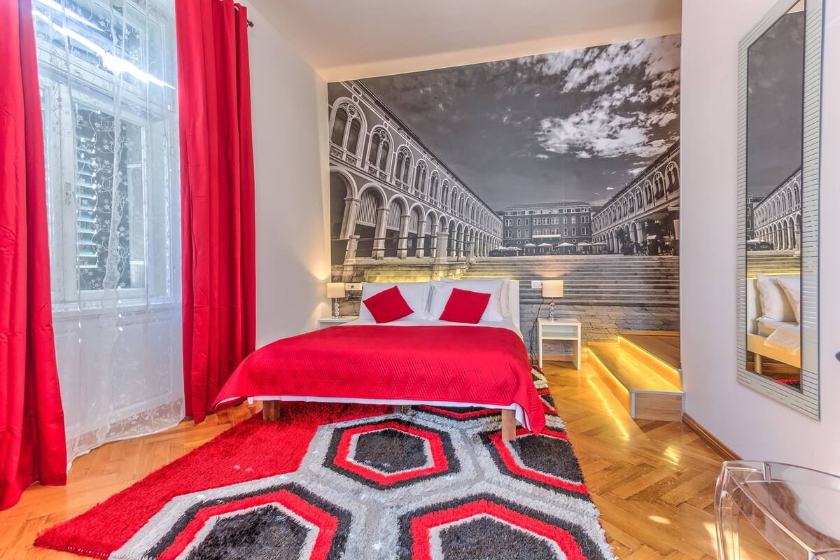 Ein Schlafzimmer mit einem großen Bett, roter Bettwäsche, einem geometrischen Teppich, einem Fenster mit roten Vorhängen und einem Wandgemälde, das eine Stadtansicht zeigt.