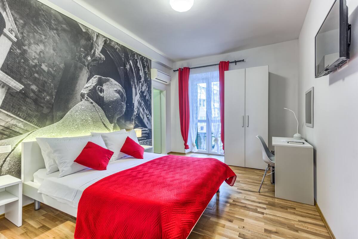 Ein modernes Schlafzimmer mit einem großen Bett, einem Schreibtisch und Blick auf die Stadt durch ein Fenster.