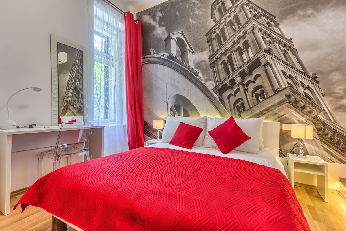 Ein Hotelzimmer mit einem großen Bett, einem Schreibtisch und einem Sessel. Ein Fenster mit roten Vorhängen und ein Wandbild eines Gebäudes sind ebenfalls zu sehen.