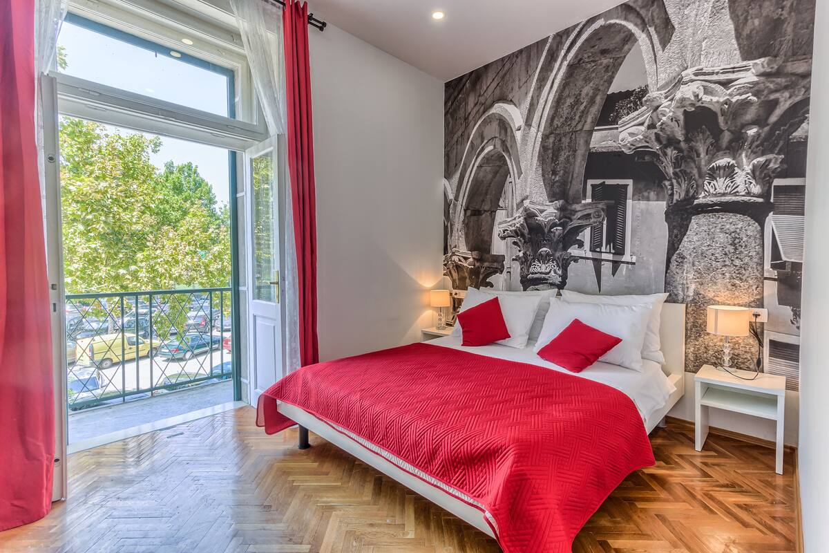 Ein Schlafzimmer mit einem großen Bett, roter Bettwäsche, Blick auf die Straße und einem Wandgemälde.
