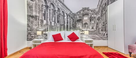Un dormitorio con una cama grande, ropa de cama roja, una alfombra roja y un mural de un edificio histórico en la pared.