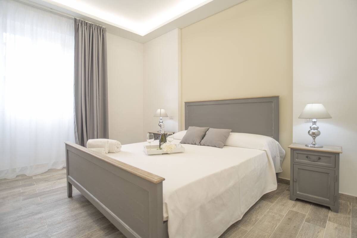 A neatly made bed with a grey headboard, a bedside table with a lamp, and a window with curtains.