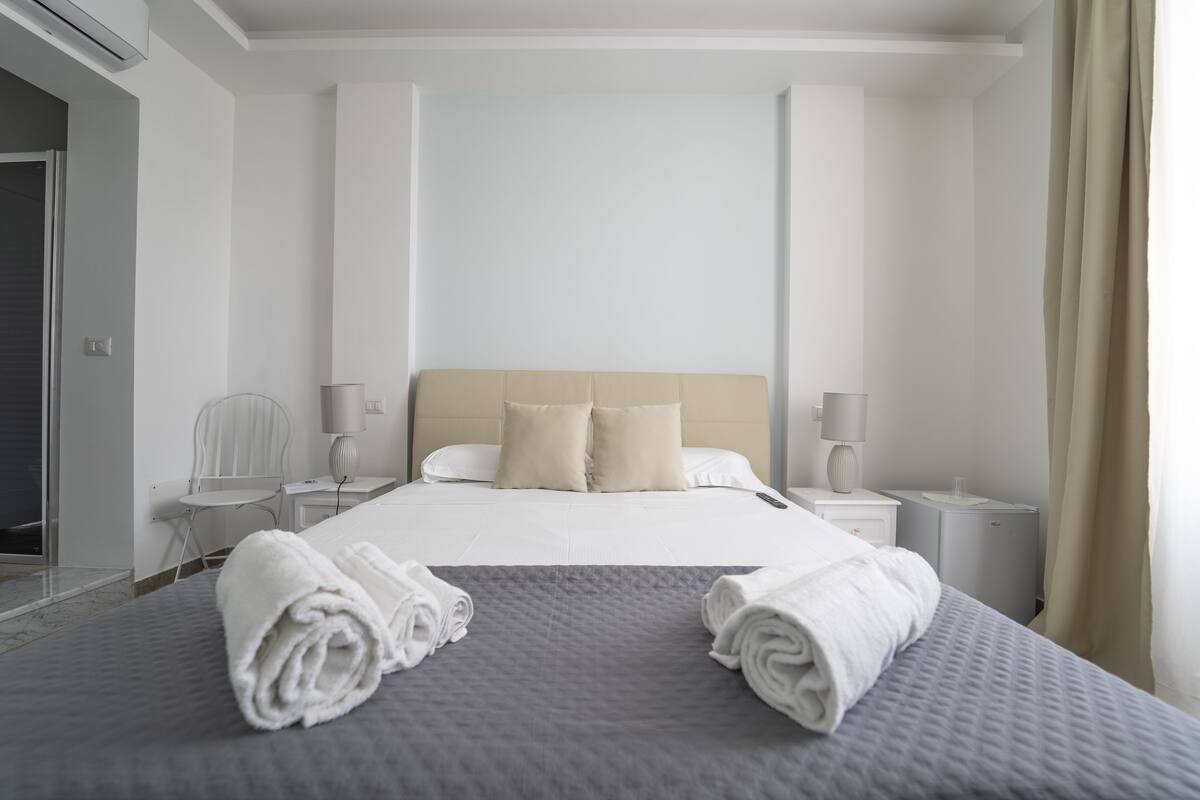 A neatly made bed with two rolled towels, a bedside table with a lamp, and a chair in a modern hotel room.