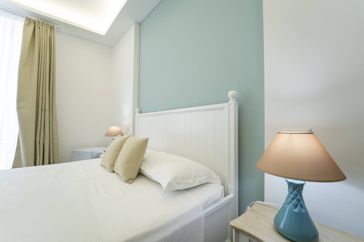 A neatly made bed with white linens and beige pillows, a bedside lamp, and a nightstand with a lamp.