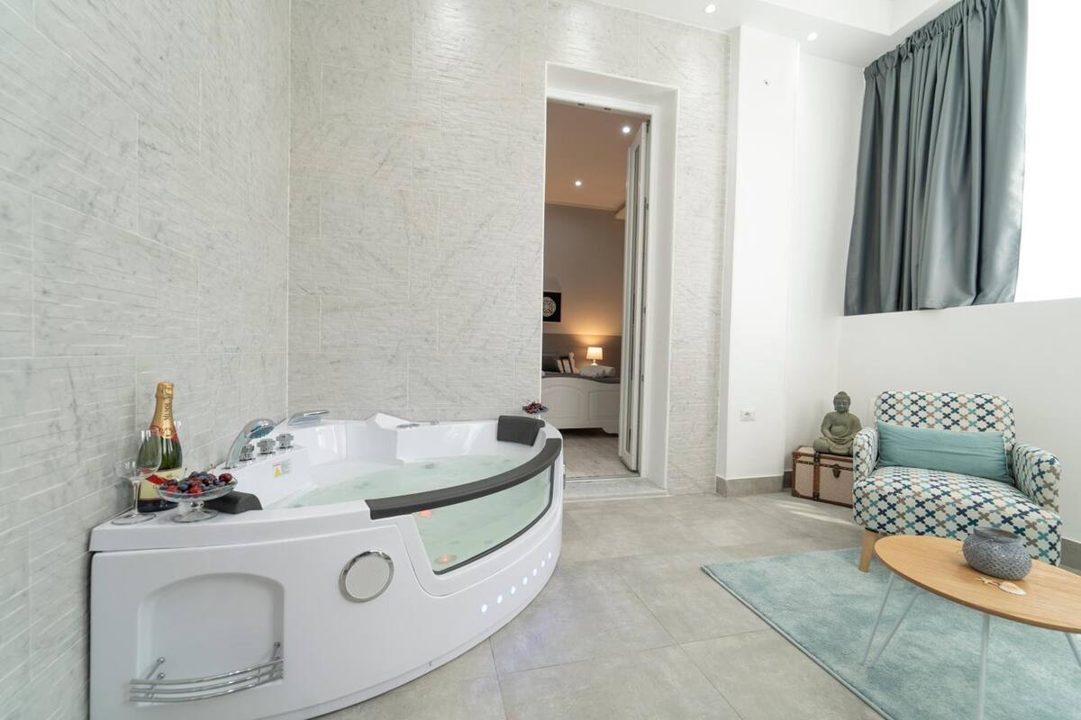 A modern bathroom with a freestanding bathtub, a small table with a bottle and fruit, and a patterned armchair.