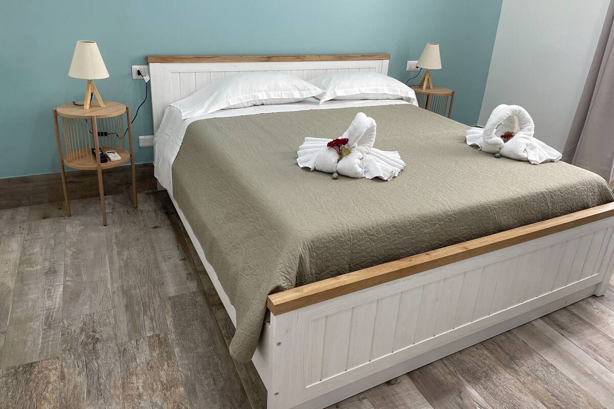 A neatly made bed with a beige bedspread, two white pillows, and two towels arranged in a decorative manner on the bed. There are two bedside tables with lamps, and the room has light blue walls and wooden flooring.