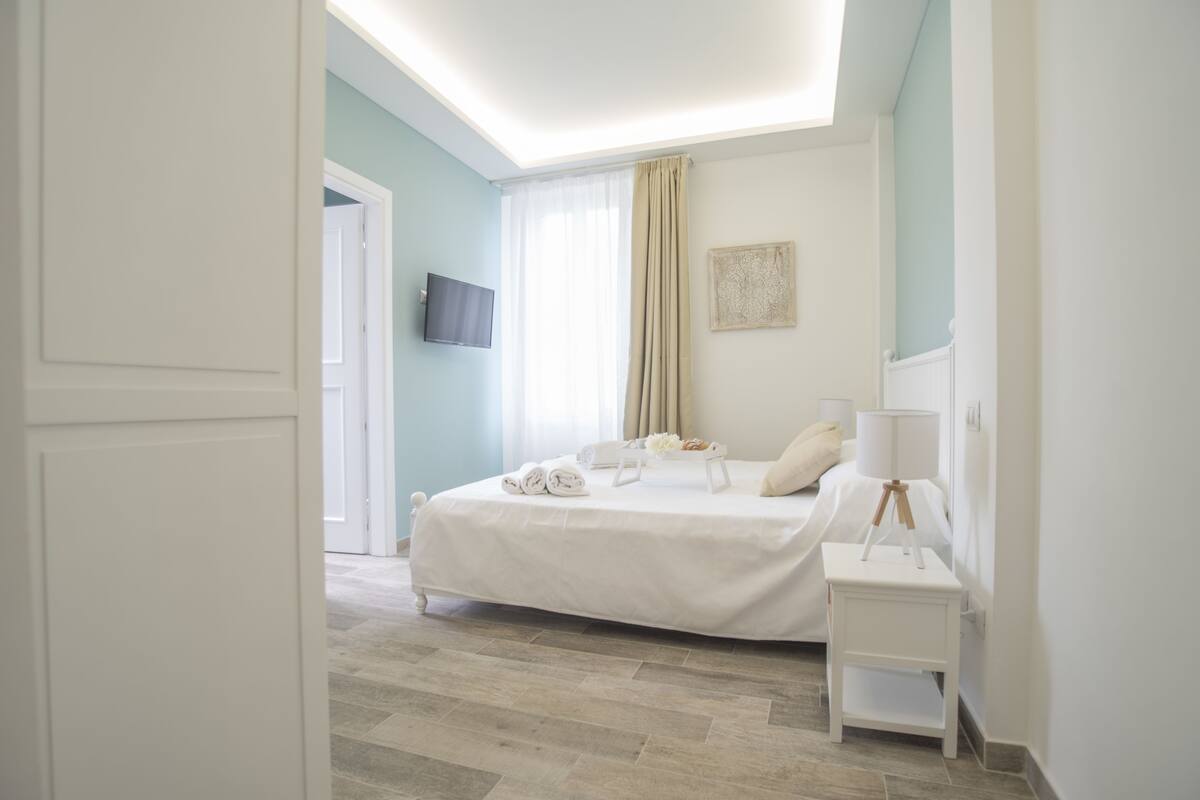 A bedroom with a bed, a bedside table, a lamp, a TV, and a window with curtains.