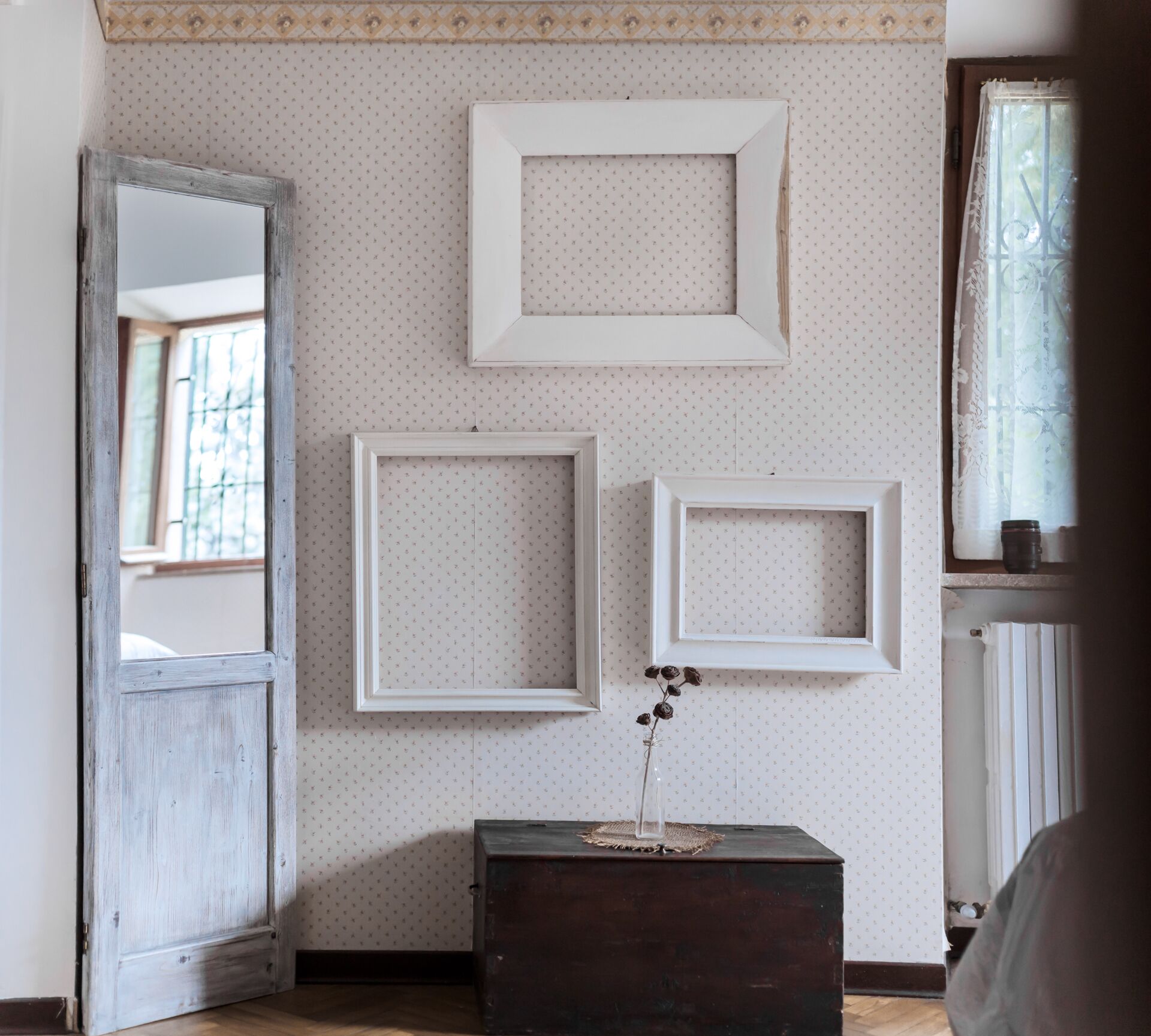 Una habitación con tres marcos blancos en la pared, una puerta de madera, una mesa de madera oscura con un jarrón de flores secas y una ventana con cortinas de encaje.