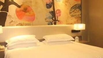 A hotel room with a bed, pillows, a nightstand, a wall-mounted lamp, and a decorative wall with abstract art.