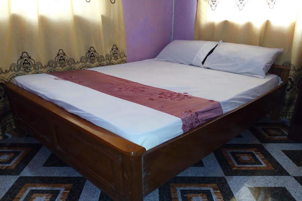 A single bed with a wooden frame, white bedding, and a red decorative strip. The room has patterned curtains and a tiled floor with geometric designs.
