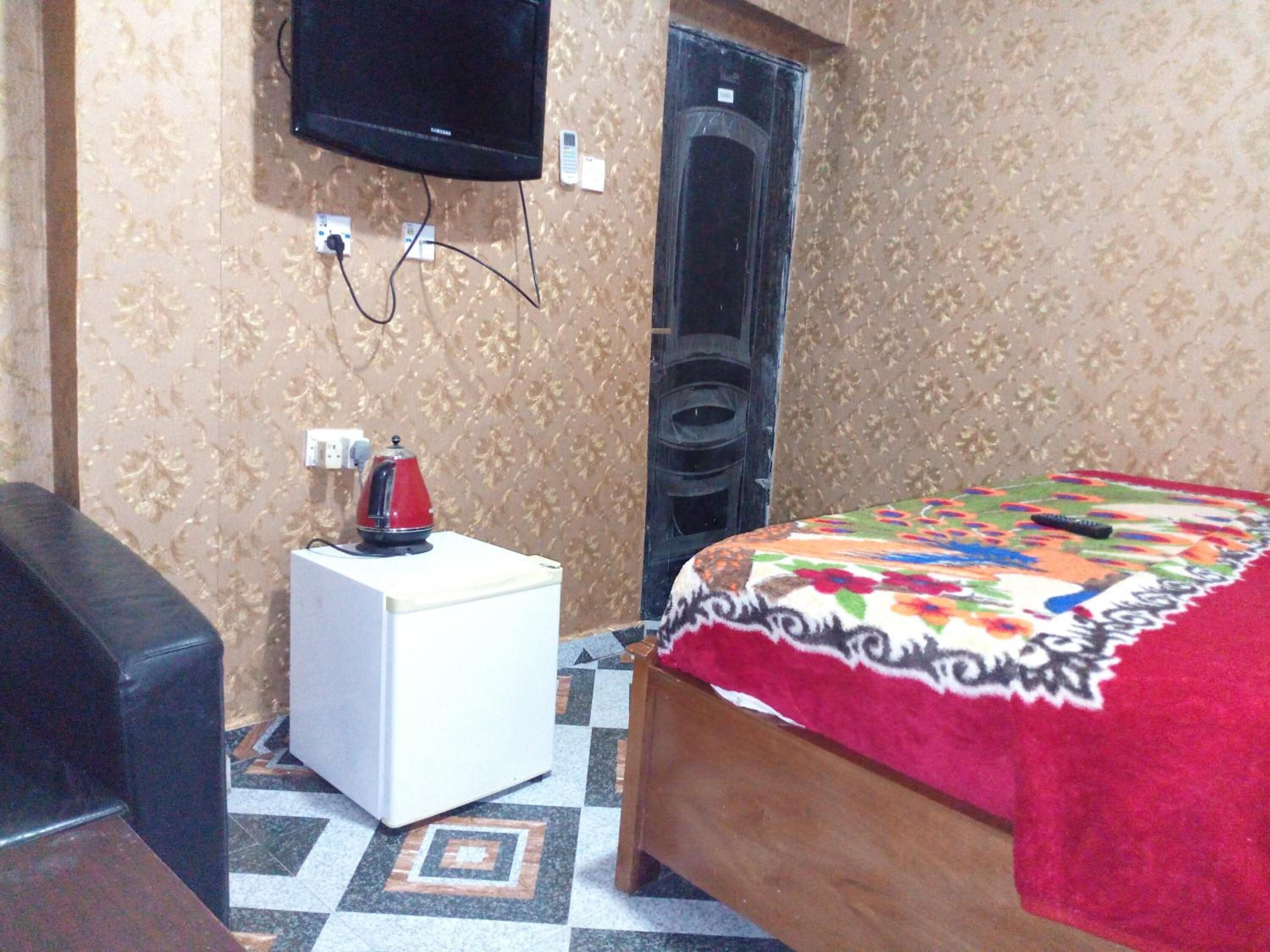 A small room with a bed, a white side table, a red kettle, a television mounted on the wall, and a patterned carpet.