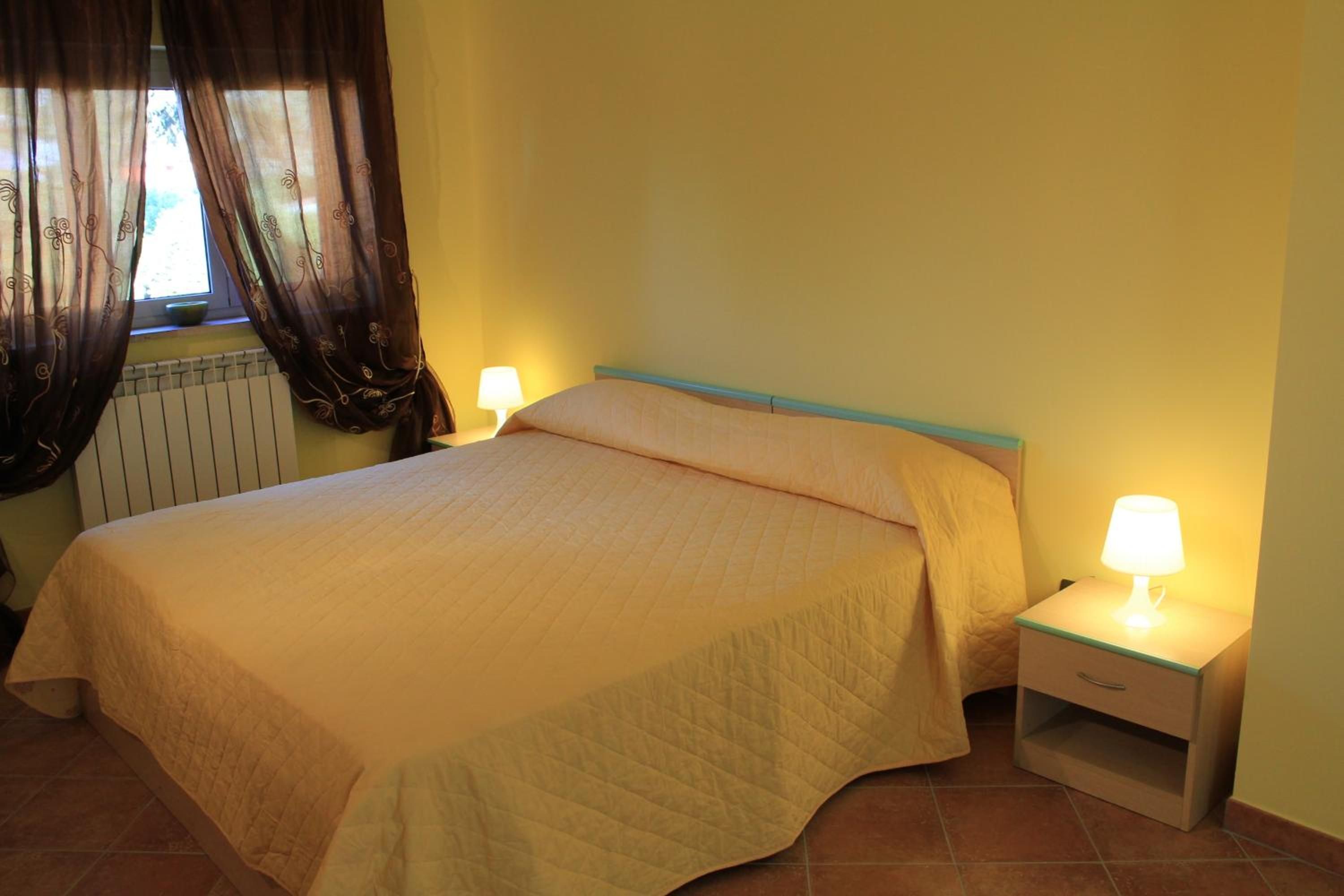 Una cama individual con colcha, dos lámparas de noche, una ventana con cortinas y un radiador.