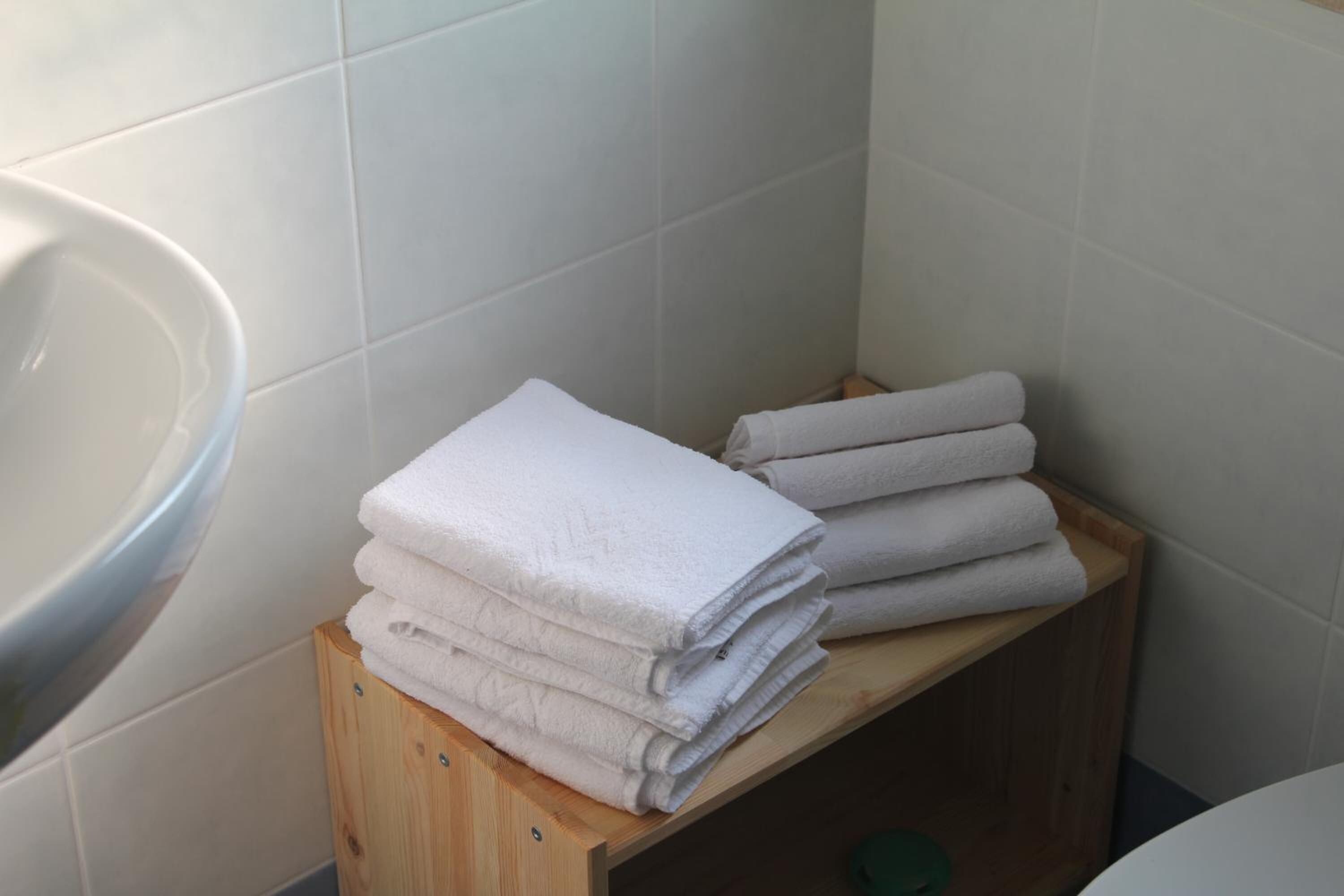 Une pile de serviettes blanches est posée sur une étagère en bois, dans une salle de bain aux carreaux blancs.