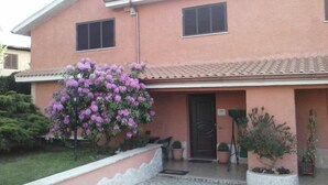 Un edificio de dos pisos con fachada rosa, techo de tejas y un pequeño porche con plantas en macetas.