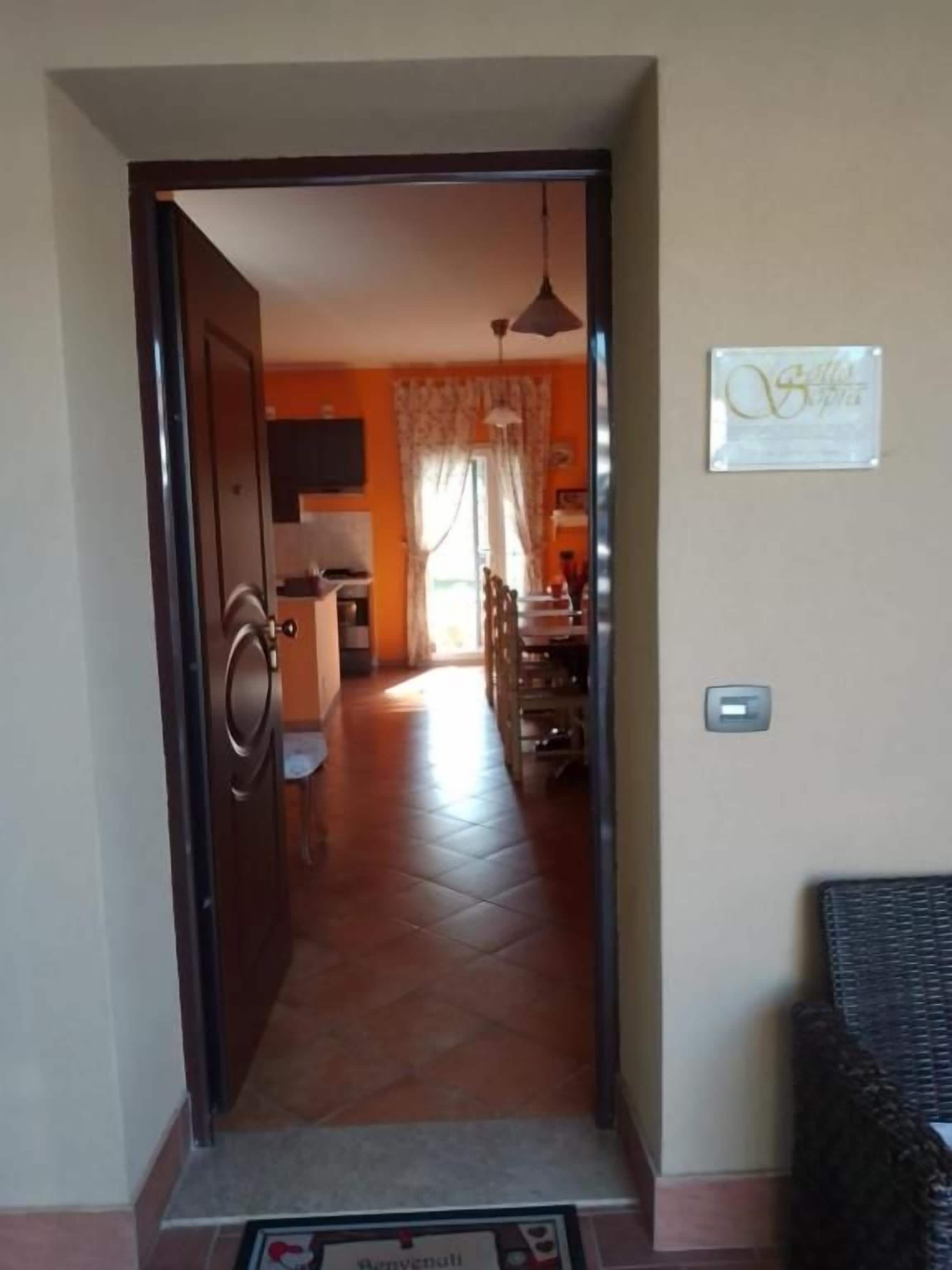Una puerta que da a un comedor con una araña, un sillón de mimbre y un felpudo.