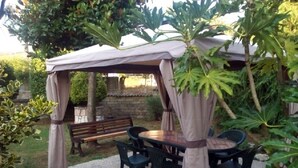 Un espacio al aire libre con mesa redonda y sillas, un banco y una pérgola con cortinas.