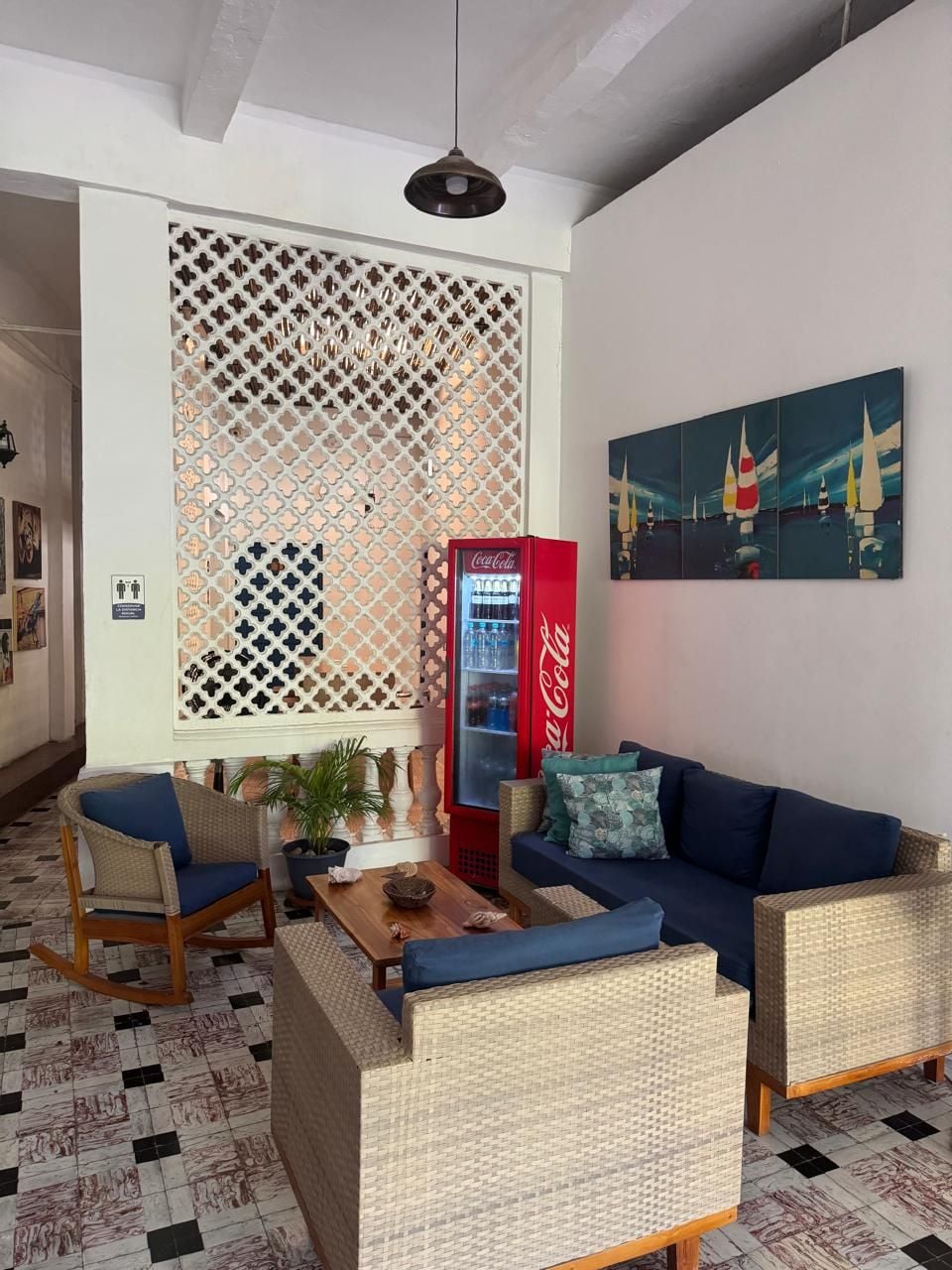 Una habitación con sofá, sillas, una mesa, un separador decorativo, un refrigerador Coca-Cola y un cuadro.