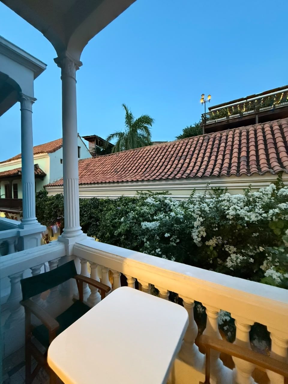 A balcony with a white table and chairs, overlooking a building with red-tiled roof and white flowering bushes.