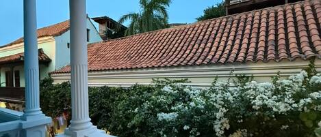A balcony with a white table and chairs, overlooking a building with red-tiled roof and white flowering bushes.