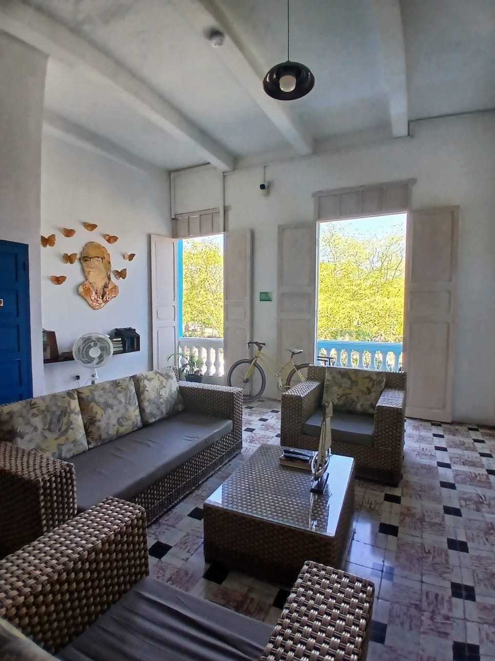 Una sala de estar con sofá, mesa de centro y ventilador. Hay dos ventanas con vistas a árboles.