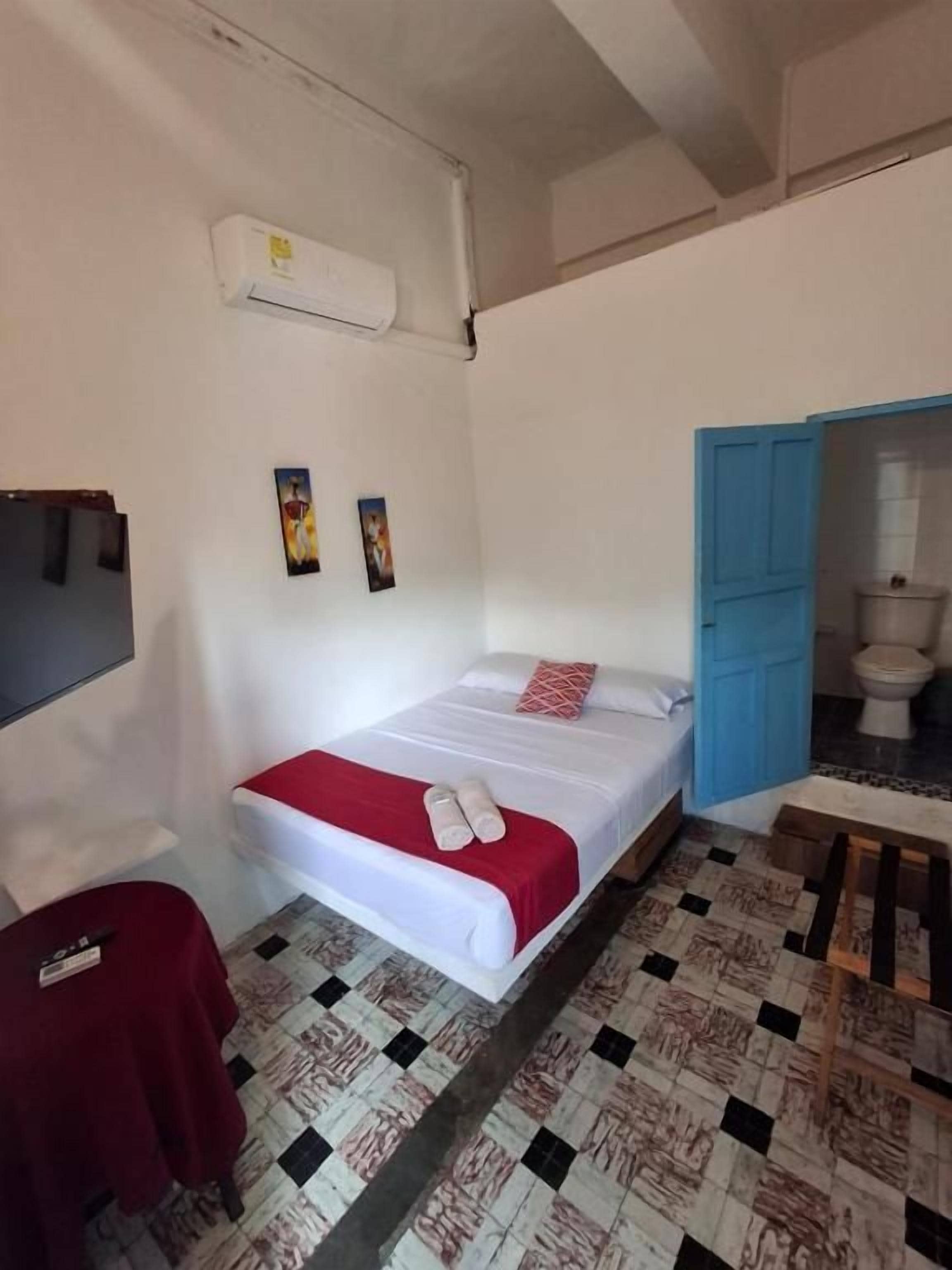 Une petite chambre avec un lit simple, un sol à motifs, une couvre-lit rouge, un climatiseur blanc fixé au mur et une porte bleue donnant sur une salle de bain.