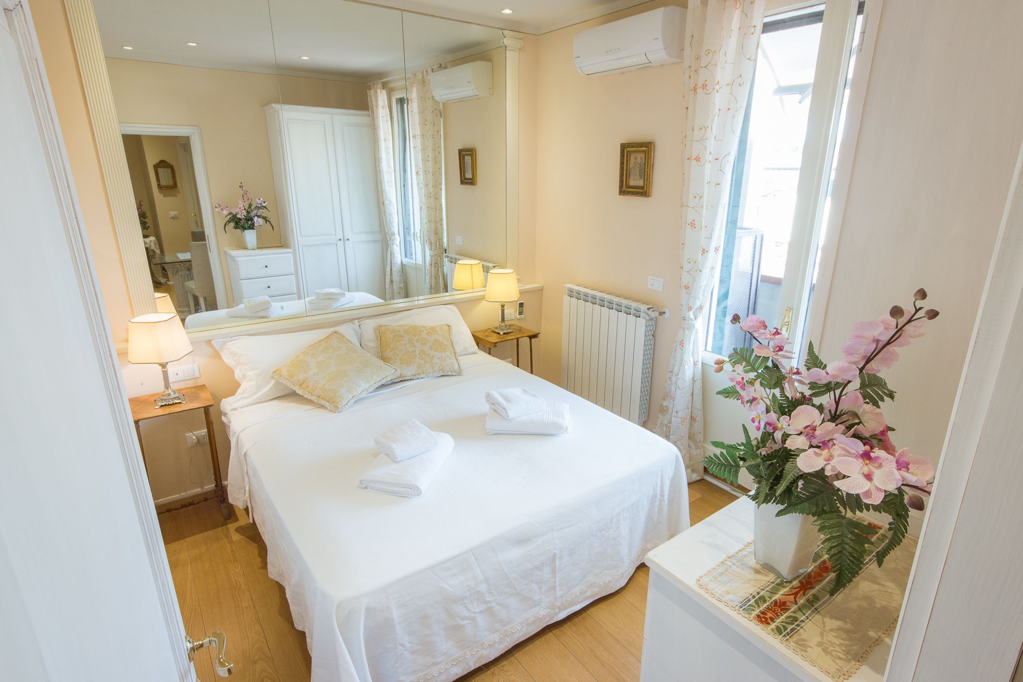 A hotel room with a bed, bedside lamps, a mirror, a radiator, and a window with curtains.