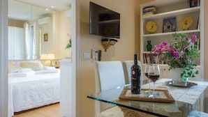 A hotel room with a bed, a glass table with a bottle and wine glass, a TV, and a bookshelf with books and decorative items. - Efeso (Florence)
