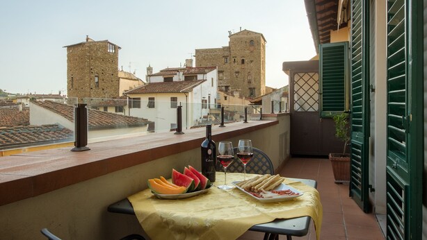 A balcony with a table set for two, featuring a bottle of wine, glasses, watermelon slices, and breadsticks. The balcony overlooks a historic town with stone buildings and a tower. - Efeso (Florence)
