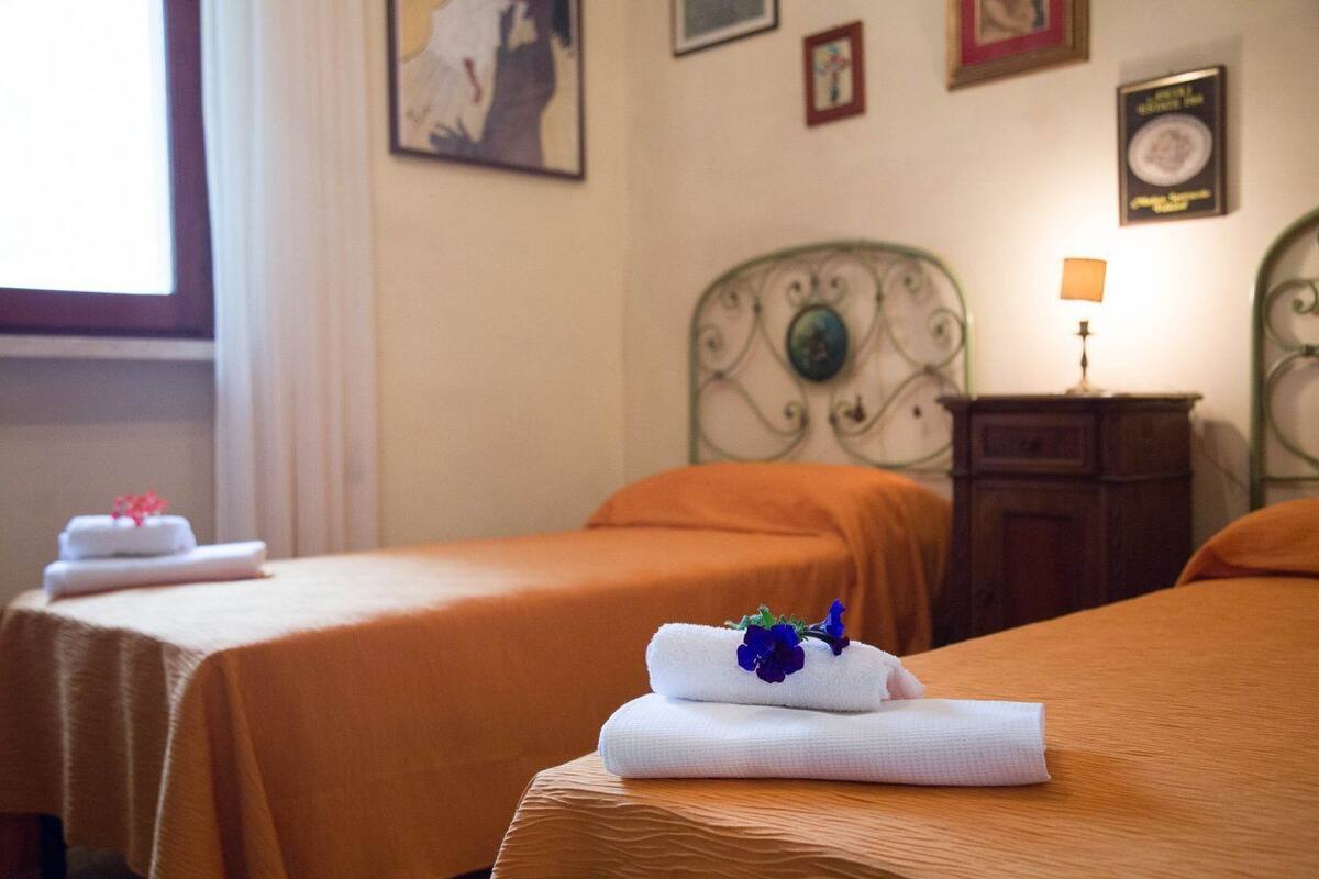 Habitación de hotel con dos camas, cada una con cubrecama naranja y toalla blanca con flor azul. Hay una mesita de noche con lámpara, un certificado enmarcado en la pared y una ventana con cortinas.