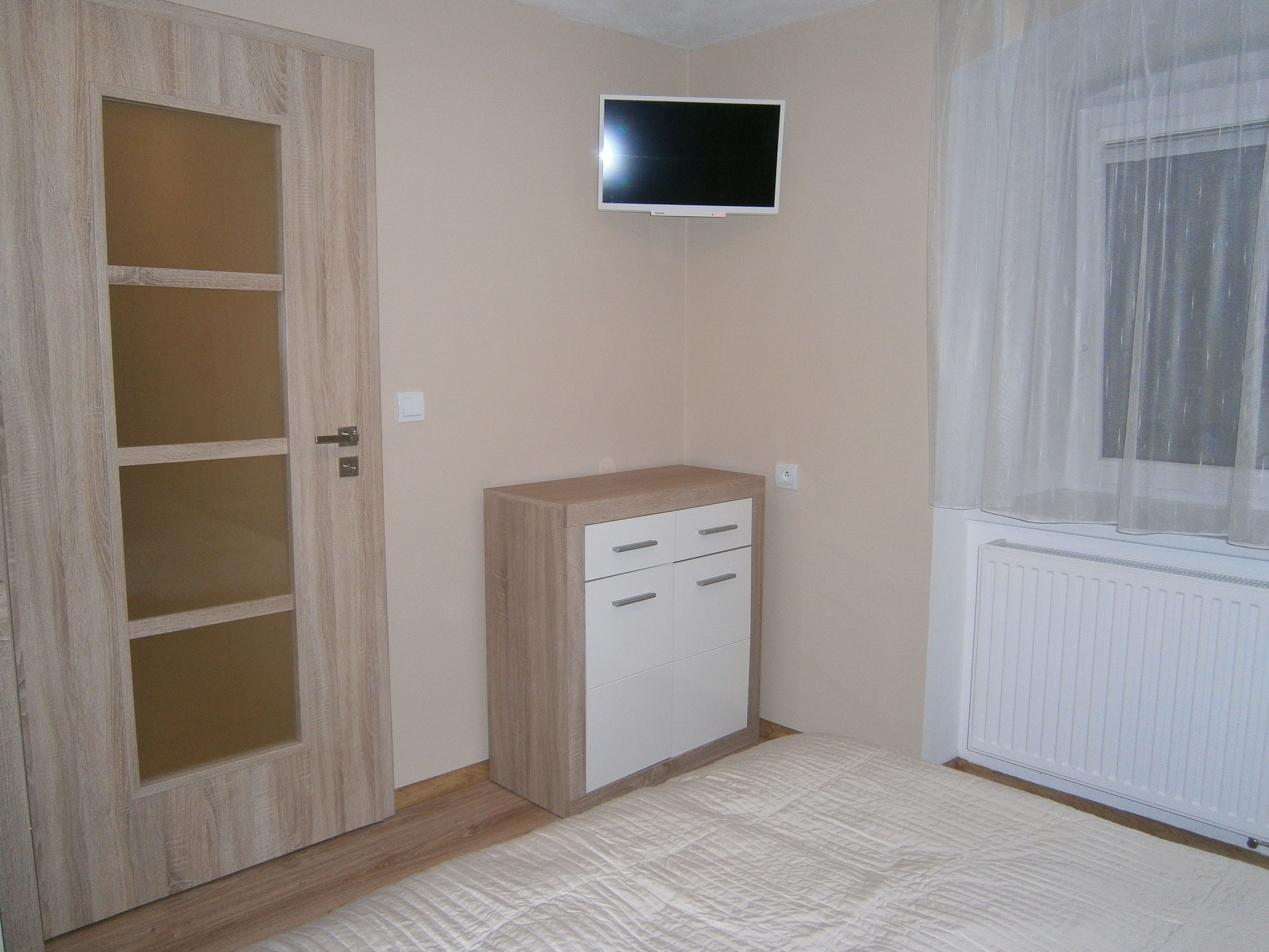 Quarto com guarda-roupa de madeira, televisão na parede e janela com cortinas leves.