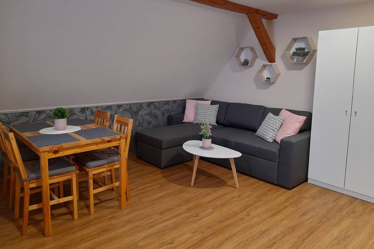 A room with a wooden dining table, chairs, a grey sofa, and a small white table with a plant.