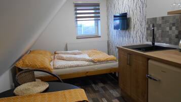 A compact room with a bed, a small table, a chair, a kitchen area with a sink, and a TV mounted on the wall.
