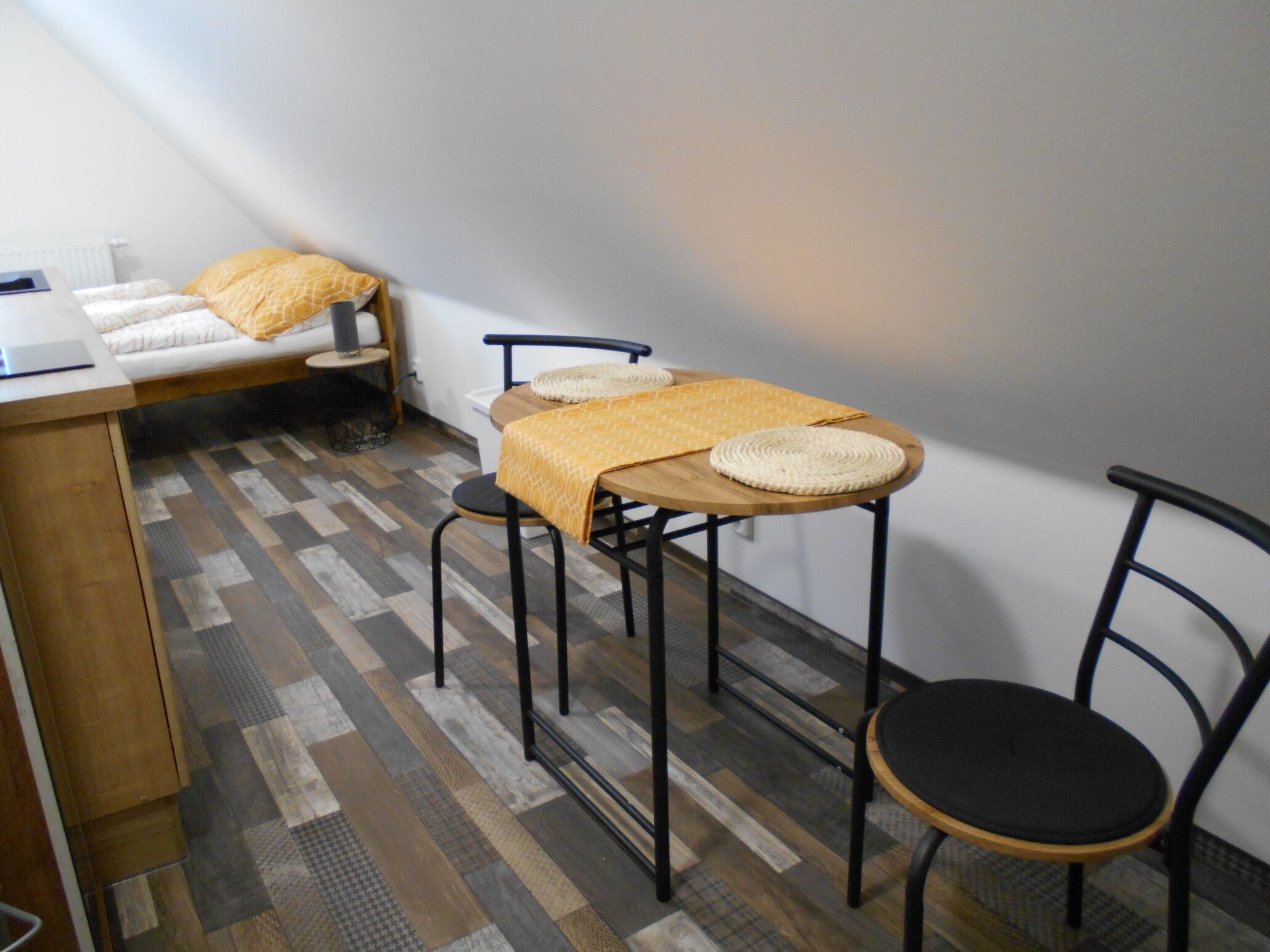 Une petite chambre moderne, avec un lit, une table en bois et deux chaises, et un sol à motifs.