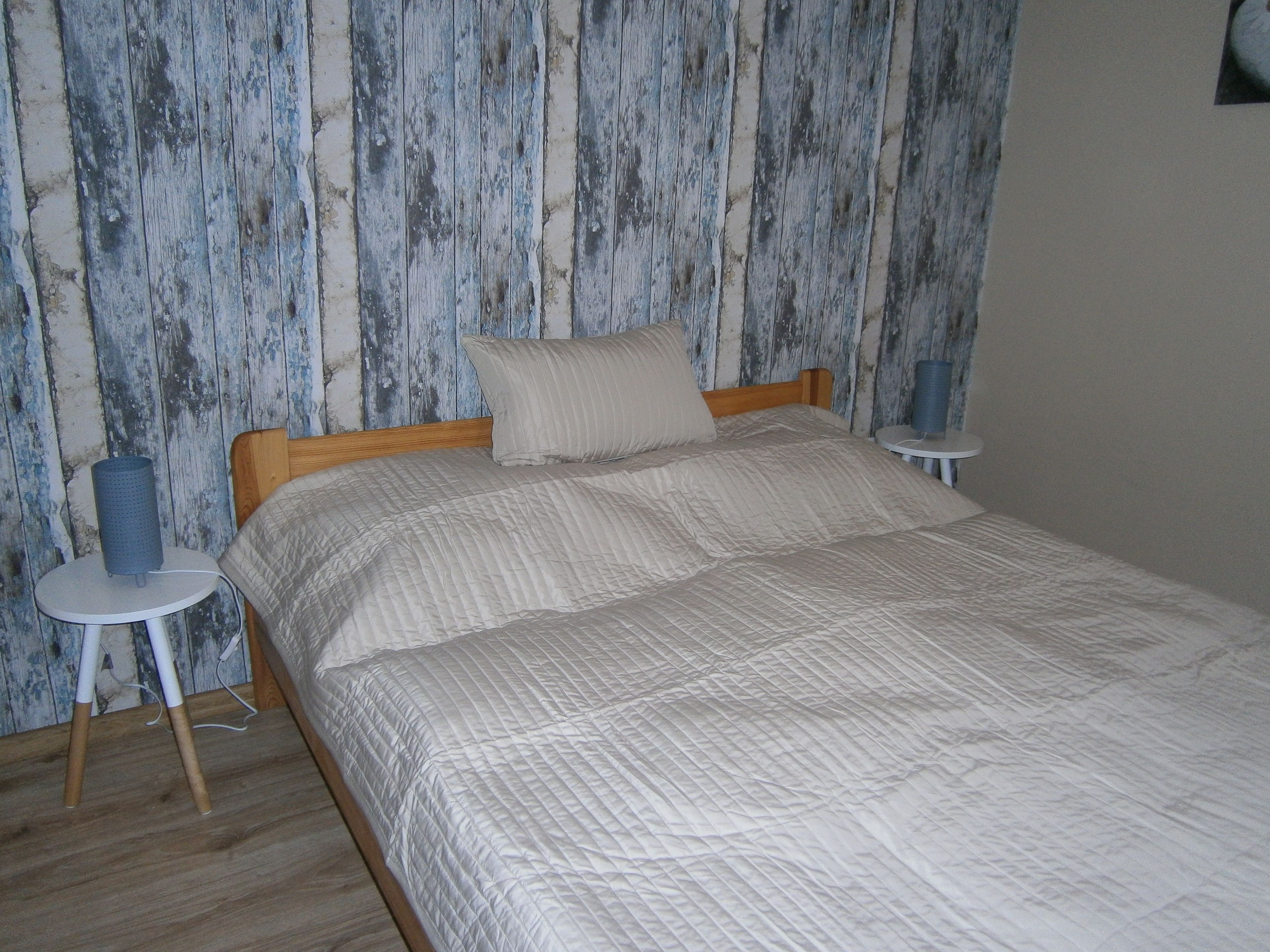 A bed with a quilted cover, a wooden headboard, and a pillow. There are two bedside tables, one with a blue cylindrical object and the other with a white object. The wall behind the bed has a distressed wood panel design.