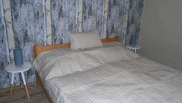 A bed with a quilted cover, a wooden headboard, and a pillow. There are two bedside tables, one with a blue cylindrical object and the other with a white object. The wall behind the bed has a distressed wood panel design.