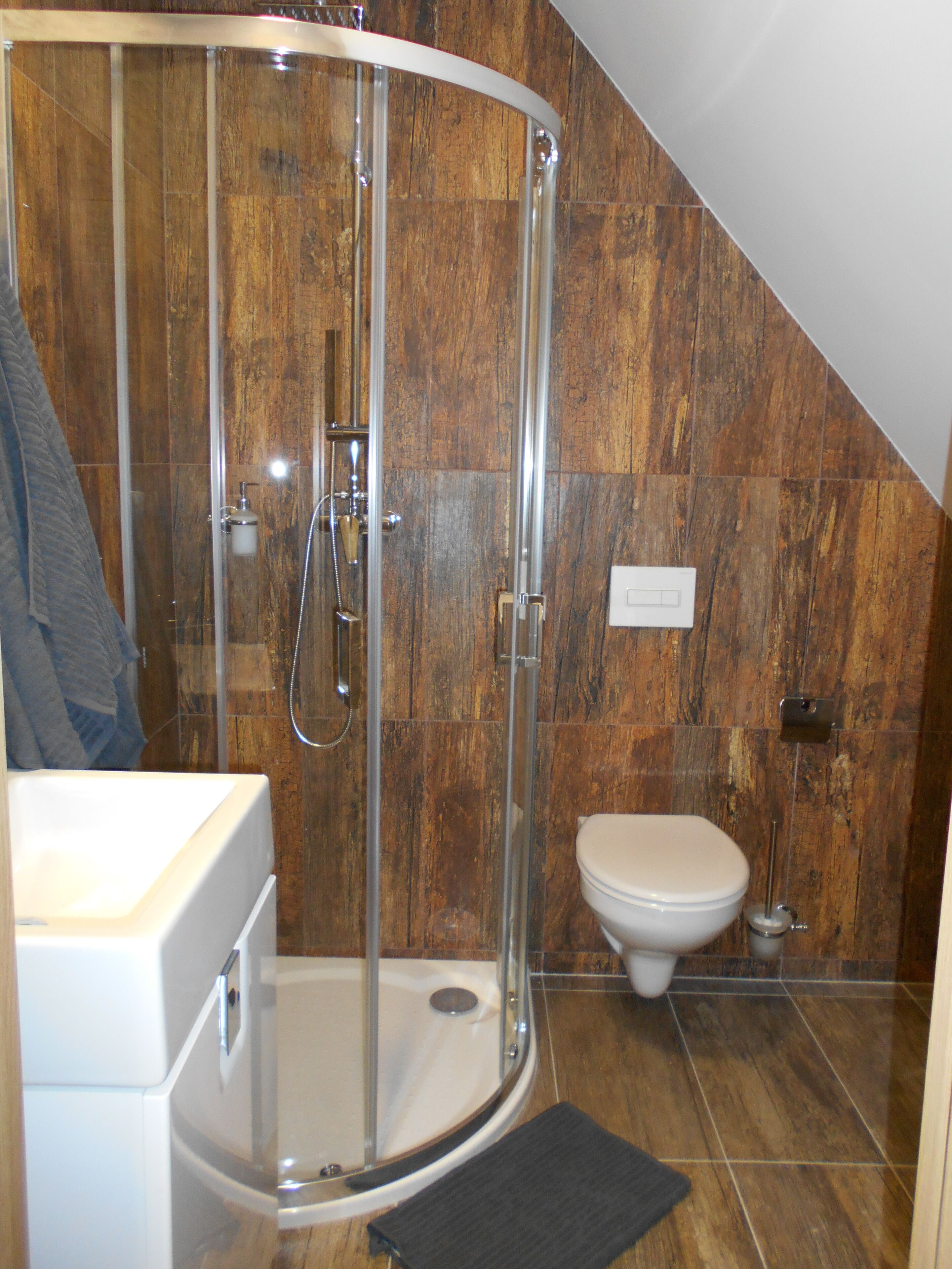 Une douche avec une porte en verre, des toilettes et un lavabo dans une salle de bain aux murs en bois.