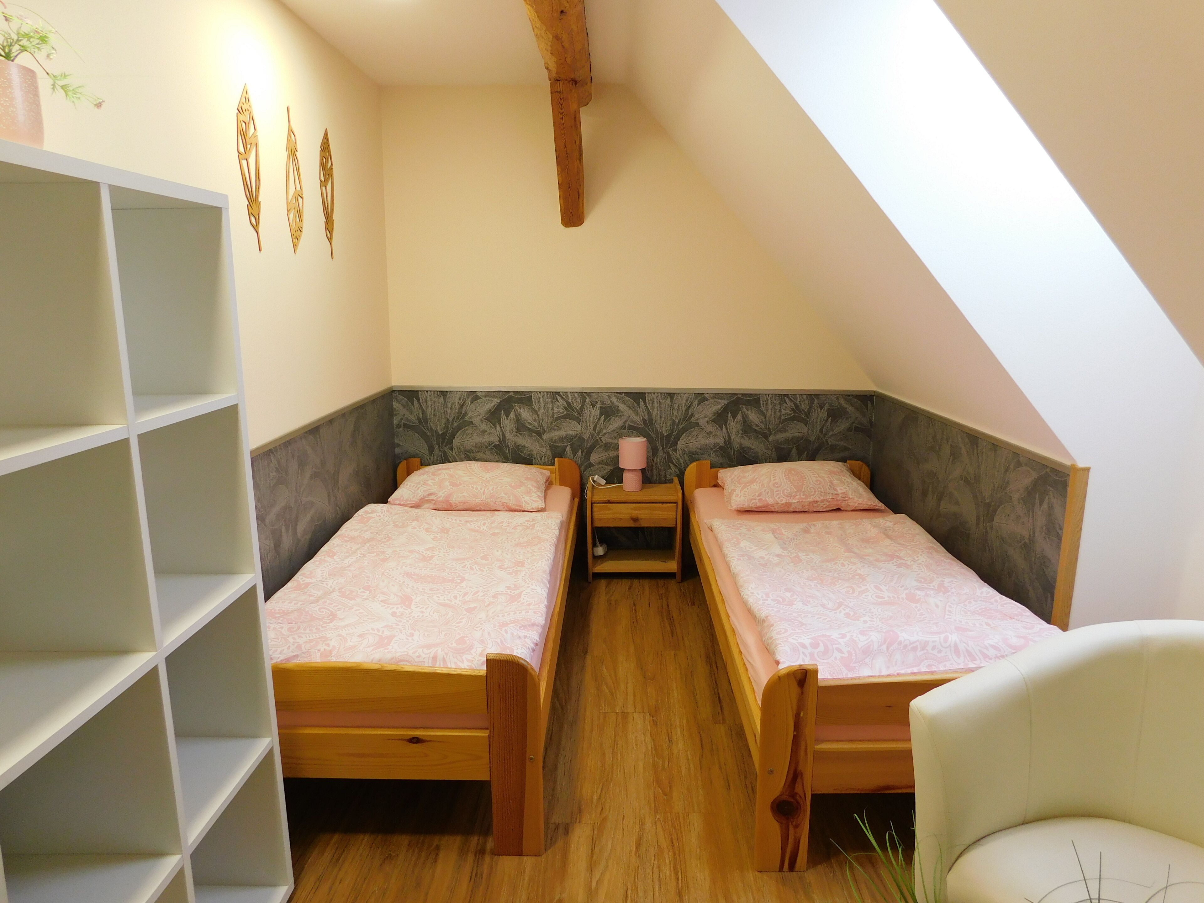 A small, cozy room with two wooden beds, a white chair, and a bookshelf.