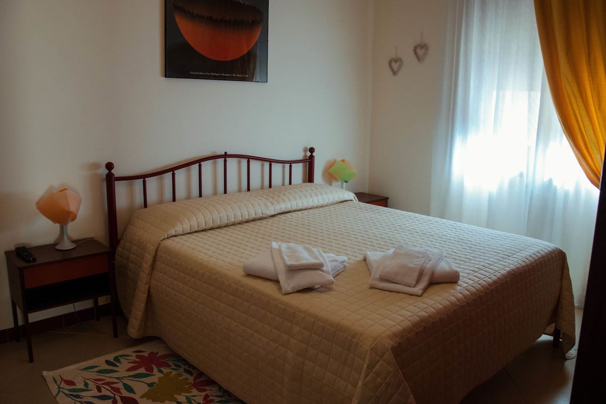 A neatly made bed with a quilted cover, a bedside table with a lamp, and a floral-patterned rug.