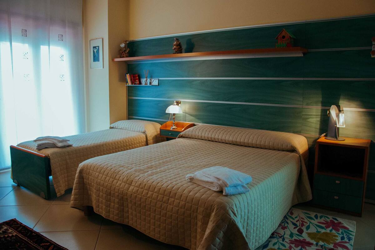 A hotel room with two beds, a nightstand with a lamp, a small shelf with decorative items, and a window with curtains.